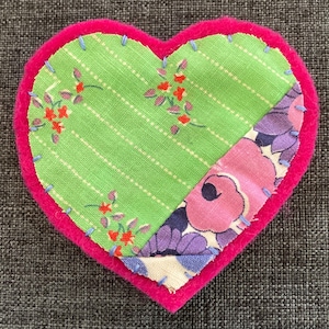 May include: A heart-shaped patch with a bright pink felt border. The interior features two fabric sections: a green striped fabric with floral accents and a floral patterned fabric in pink and purple. Light blue stitching secures the fabrics.