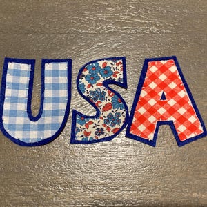 May include: Three fabric letters spelling USA, each outlined in dark blue felt with white stitching. The U features a light blue gingham pattern, the S displays a floral print with blue and red flowers, and the A has a red and white gingham pattern.
