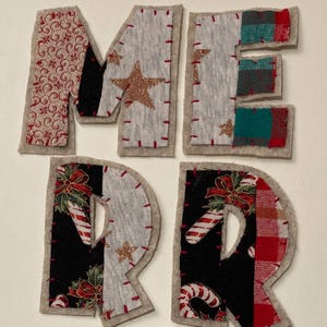 May include: Four fabric letters spelling M, E, R, R, constructed from beige felt backing with various patterned fabric patches including red swirls, grey with gold stars, plaid, and holiday prints featuring candy canes and holly, all joined by red stitching.