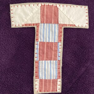 Pode incluir: Uma letra T de patchwork feita de tecido listrado vermelho e branco e tecido branco. A letra é costurada com fio vermelho e tem um suporte bege.