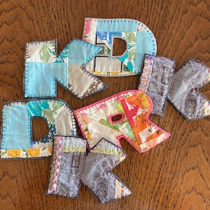 Peut inclure: Ensemble de lettres en tissu épelant "DRK". Chaque lettre est un patchwork de divers tissus à motifs dans des tons de bleu, rose, orange et gris, avec des coutures visibles. Les lettres sont disposées sur une surface en bois.