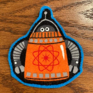 May include: A fabric patch with a cartoon robot design. The robot has an orange body with a red atomic symbol, grey arms, and a black head. The patch is outlined in blue.