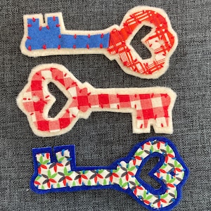 Könnte beinhalten: Drei dekorative Schlüssel-förmige Stoffaufnäher. Einer ist blau mit roten Nähten, ein anderer rot und weiß kariert, und der dritte ist blau mit einem Blumenmuster. Alle haben einen weißen Rand.