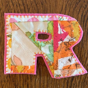 Może przedstawiać: Ręcznie wykonana litera "R" z tkaniny z różową filcową obwódką. Litera składa się z różnych wzorzystych tkanin w odcieniach pomarańczu, zieleni i różu, zszytych pomarańczową nicią. Litera ma wycięty fragment.