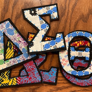 以下が含まれることがあります： 黒い縁取りのある、カラフルな布製の文字のコレクション。文字は、花柄、幾何学模様、抽象的なデザインなど、さまざまな模様の生地で作られています。各文字は個別にカットされ、縫い付けられており、パッチワーク効果を生み出しています。文字は木製の表面に配置されています。