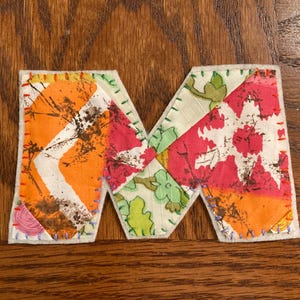 May include: A patchwork fabric letter 'M' with a colourful design. The letter is made up of sections of orange, pink, and green floral patterns, stitched together with coloured thread. The edges are finished with a white felt backing.