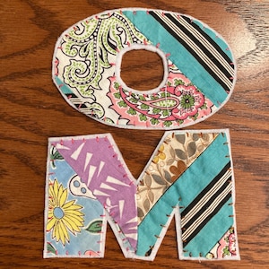 Könnte beinhalten: Ein handgefertigter Stoffbuchstabe "O" und ein handgefertigter Stoffbuchstabe "M" im Patchwork-Stil. Die Buchstaben bestehen aus verschiedenen Stoffen mit floralen, Paisley- und gestreiften Mustern in den Farben Rosa, Blau, Grün und Weiß. Die Buchstaben sind mit rosa Garn zusammengenäht und haben einen weißen Rand.