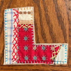 May include: A handcrafted, patchwork letter "L" composed of various fabric scraps. The design incorporates red floral patterns, blue gingham, and cream-colored fabrics, all stitched together with red, blue, and yellow thread. The letter is set against a wooden background.