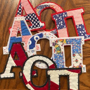 May include: A collection of fabric letters in various colors and patterns, including red, blue, and floral prints. The letters are stitched onto a white felt backing, creating a patchwork design. The letters are arranged on a wooden surface.