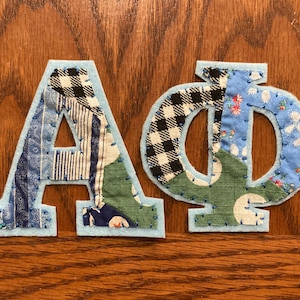 Op de afbeelding: Opgeplakte letters A, O en Φ met een lichtblauwe viltrand. De letters zijn gemaakt van stofresten in verschillende patronen, waaronder blauwe paisley, zwart-wit ruitjes en bloemenprints. De letters staan op een houten oppervlak.