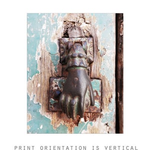 May include: A dark metal door knocker shaped like a hand, mounted on a weathered wooden door with peeling light green paint. The knocker features a cuff detail at the wrist and is attached to a rectangular metal plate showing signs of oxidation.