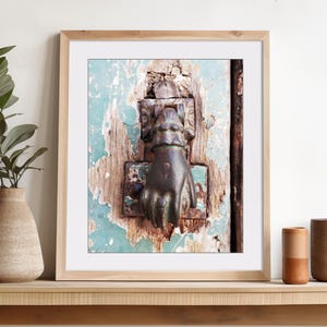 May include: A framed wall art print featuring a close-up of a dark metal door knocker shaped like a hand, mounted on a weathered, light blue painted wooden door with visible wood grain and peeling paint textures, displayed in a light wood frame.