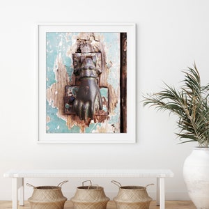 May include: A framed wall art print featuring a weathered, teal-painted wooden door with a dark, metallic hand-shaped door knocker. The door surface shows significant paint chipping and wood texture, captured in a vertical orientation.