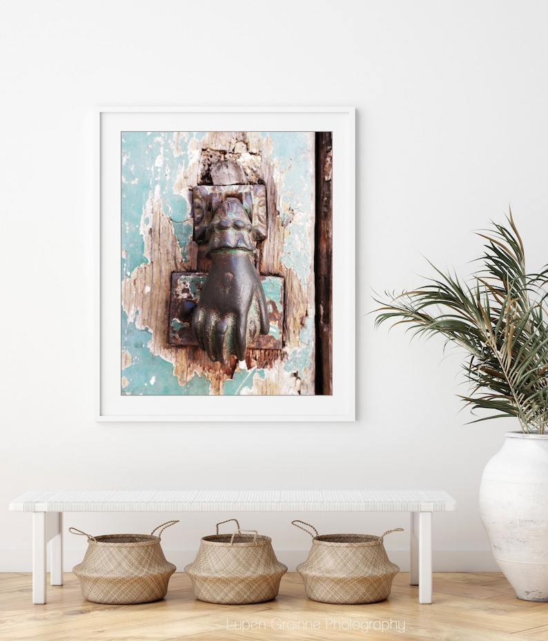 May include: A framed wall art print featuring a weathered, teal-painted wooden door with a dark, metallic hand-shaped door knocker. The door surface shows significant paint chipping and wood texture, captured in a vertical orientation.