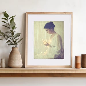 May include: A framed art print featuring a woman holding glowing lights. The artwork is in a wooden frame with a white mat. A vase with greenery and two small vases are on a wooden shelf, along with two ceramic cylinders.