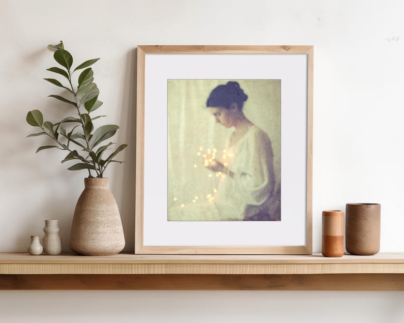 May include: A framed art print featuring a woman holding glowing lights. The artwork is in a wooden frame with a white mat. A vase with greenery and two small vases are on a wooden shelf, along with two ceramic cylinders.
