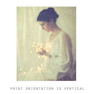 May include: A vertical print with a soft, vintage aesthetic. The image features a blurred figure holding glowing fairy lights. The figure is wearing a white garment. The text at the bottom reads "PRINT ORIENTATION IS VERTICAL."