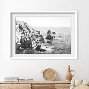 May include: A black and white framed landscape photograph depicting a rugged coastline with large rock formations rising from the ocean. The scene features a steep cliffside on the left and scattered sea stacks extending into the calm water.