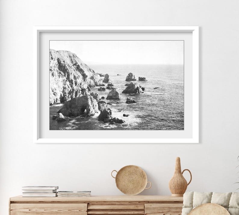 May include: A black and white framed landscape photograph depicting a rugged coastline with large rock formations rising from the ocean. The scene features a steep cliffside on the left and scattered sea stacks extending into the calm water.
