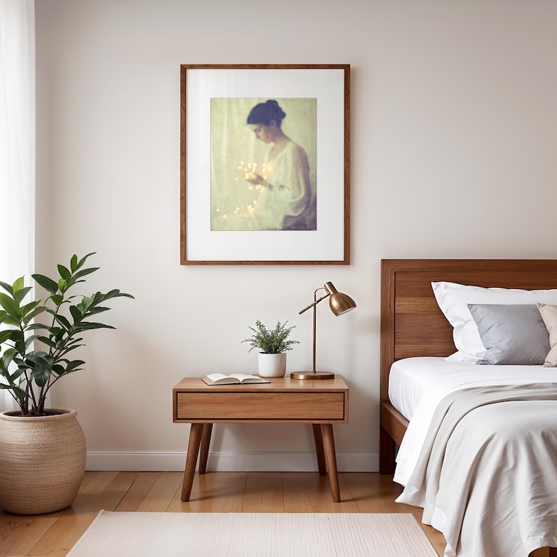 May include: A framed artwork depicts a person holding glowing lights. The artwork hangs above a wooden nightstand with a drawer, a small plant, and a brass lamp. A bed with a wooden headboard and white bedding is visible.