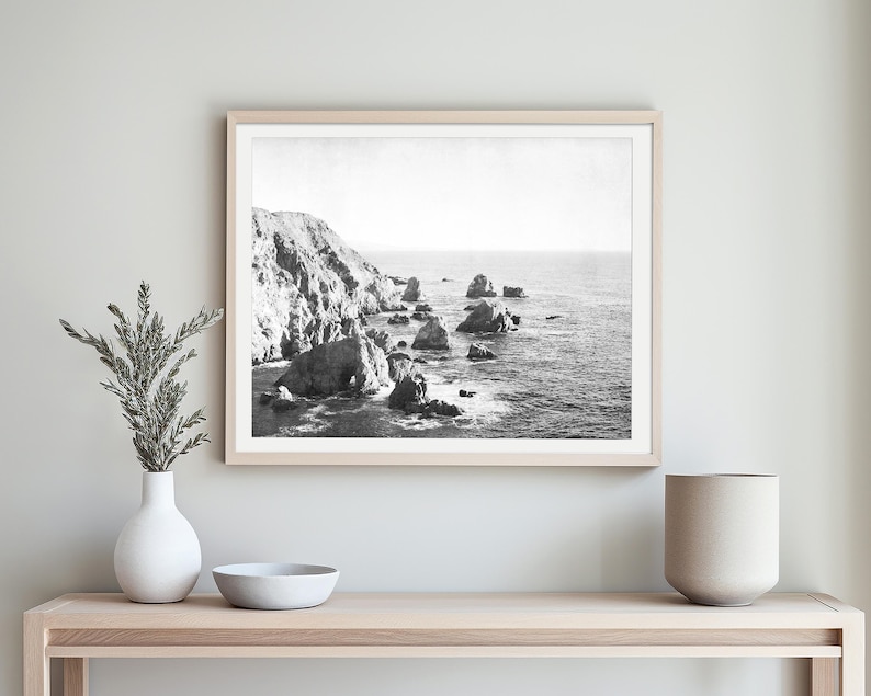 May include: A black and white landscape photograph in a light wood frame depicting a rugged coastline with large rock formations rising from the ocean. The scene captures a steep cliffside meeting the sea with scattered rocks in the water.