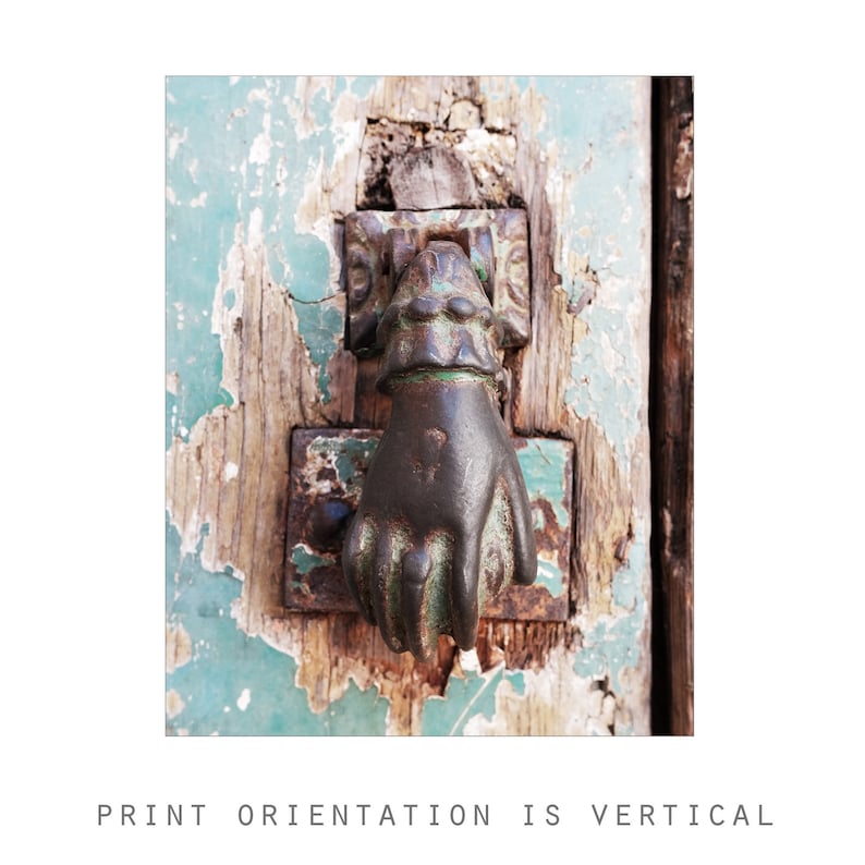 May include: A dark metal door knocker shaped like a hand, mounted on a weathered wooden door with peeling light green paint. The knocker features a cuff detail at the wrist and is attached to a rectangular metal plate showing signs of oxidation.