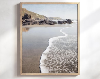 May include: A framed wall art print featuring a coastal landscape with dark rocky cliffs meeting a sandy beach. A thin line of white sea foam from a gentle wave curves across the wet sand, reflecting the soft light of a pale sky.