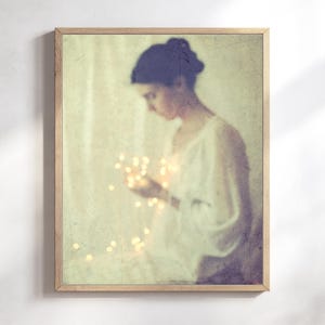 May include: A framed art print featuring a soft, vintage-style image of a person holding glowing lights. The person is wearing a white garment, and the overall color palette is muted with warm tones. The wooden frame adds a classic touch.