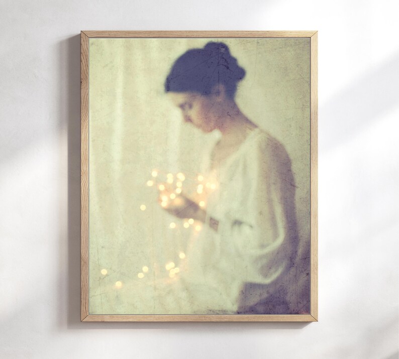 May include: A framed art print featuring a soft, vintage-style image of a person holding glowing lights. The person is wearing a white garment, and the overall color palette is muted with warm tones. The wooden frame adds a classic touch.