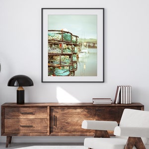 May include: A framed print featuring a coastal scene. The artwork showcases stacked crab traps with turquoise and white rope, set against a backdrop of a calm harbor and a white boat. The image has a muted, vintage aesthetic.