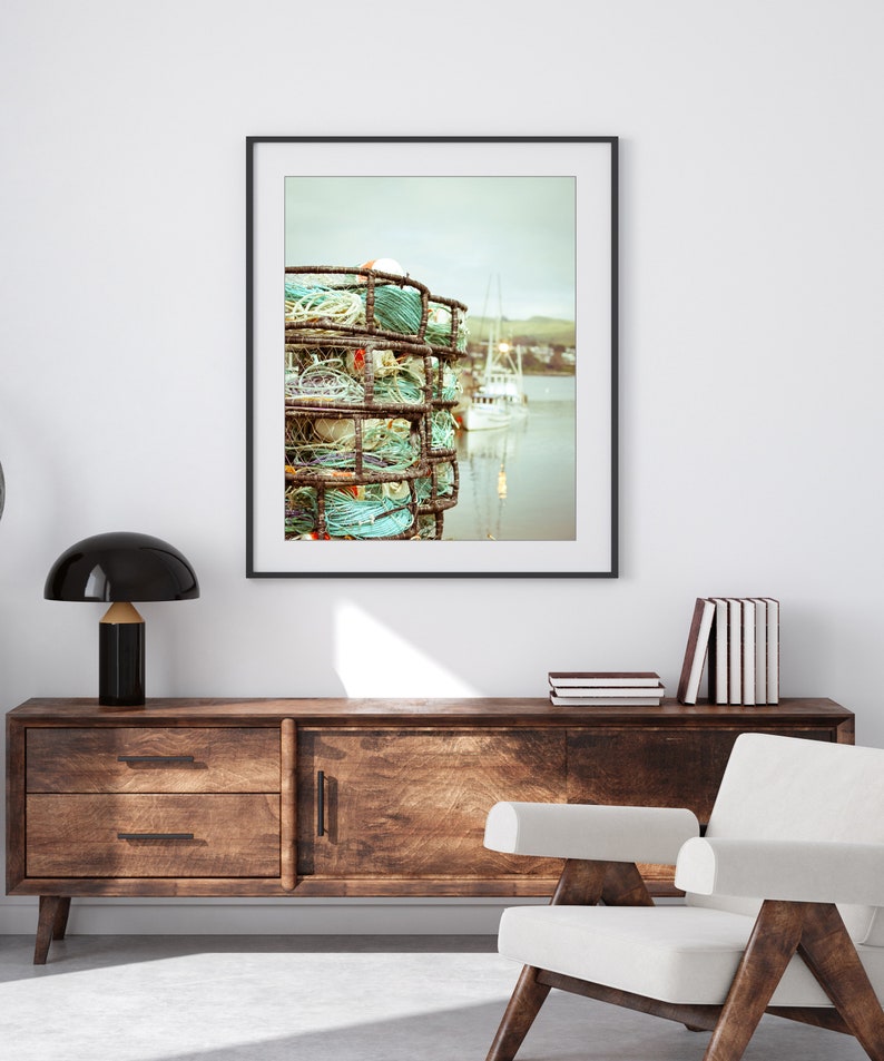 May include: A framed print featuring a coastal scene. The artwork showcases stacked crab traps with turquoise and white rope, set against a backdrop of a calm harbor and a white boat. The image has a muted, vintage aesthetic.