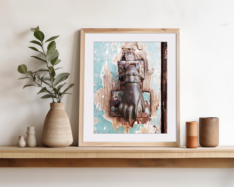 May include: A framed wall art print featuring a close-up of a dark metal door knocker shaped like a hand, mounted on a weathered, light blue painted wooden door with visible wood grain and peeling paint textures, displayed in a light wood frame.
