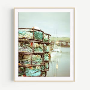 May include: A framed photograph of a crab trap filled with coiled rope in shades of blue, green, and white. A boat is visible in the background, with a soft focus effect. The image has a vintage, nautical theme.