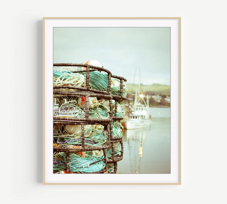 May include: A framed photograph of a crab trap filled with coiled rope in shades of blue, green, and white. A boat is visible in the background, with a soft focus effect. The image has a vintage, nautical theme.