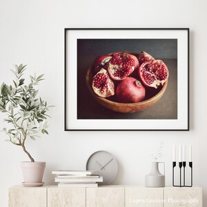 May include: A framed wall art print featuring a wooden bowl filled with whole and halved pomegranates displaying deep red seeds. The artwork is displayed in a black frame above a light wood console table with a small potted plant and candles.