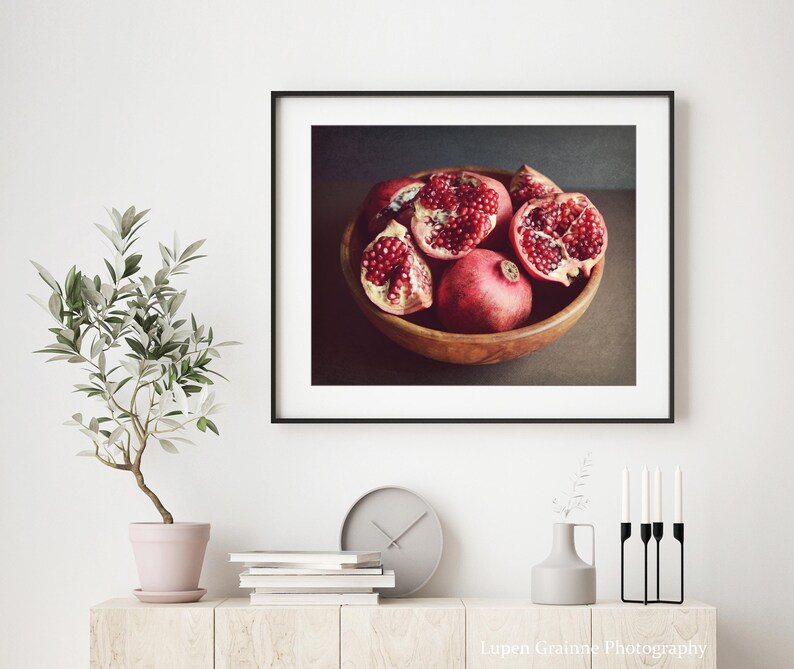 May include: A framed wall art print featuring a wooden bowl filled with whole and halved pomegranates displaying deep red seeds. The artwork is displayed in a black frame above a light wood console table with a small potted plant and candles.
