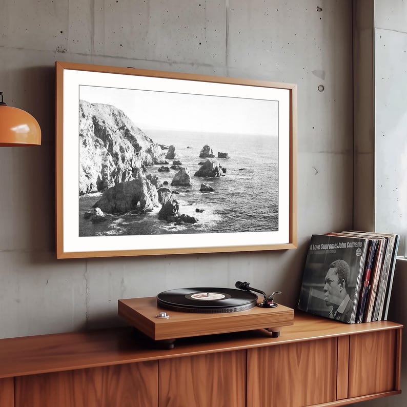 May include: A framed black and white landscape photograph of a rocky coastline with ocean waves, mounted on a concrete wall above a wooden record player and a stack of vinyl records, including a John Coltrane A Love Supreme album cover.