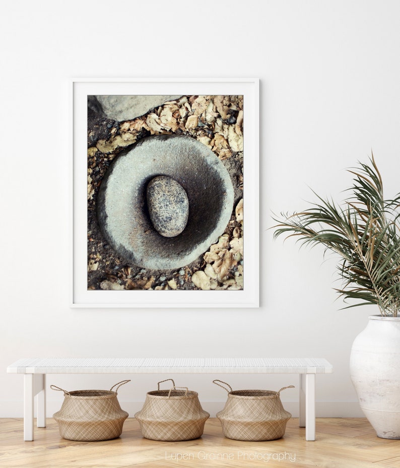 May include: A framed wall art print featuring a top-down view of a circular stone mortar with a smooth, speckled oval pestle resting inside. The stone is surrounded by dry, fallen brown leaves and dark soil, captured in a high-contrast, natural outdoor setting.