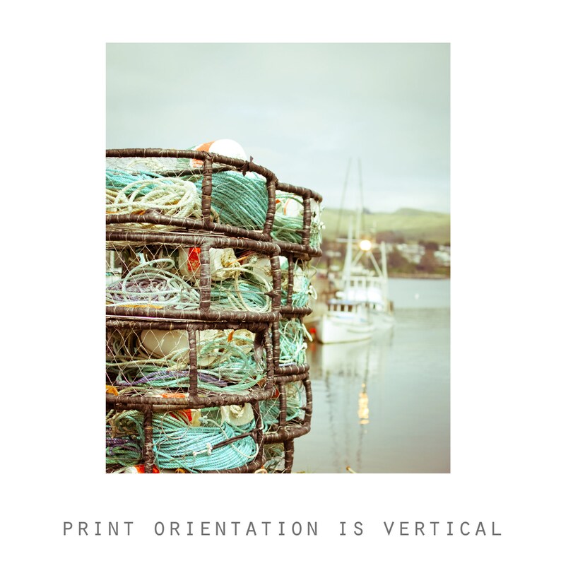 May include: A stack of crab traps filled with coiled ropes and buoys. The traps are made of dark metal and are filled with turquoise, white, and cream-colored ropes. A boat is visible in the background. The text at the bottom reads: PRINT ORIENTATION IS VERTICAL.