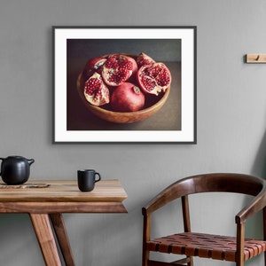 May include: A framed wall art print featuring a wooden bowl filled with several whole and halved pomegranates showing deep red seeds. The artwork is displayed in a dark frame, showcasing a still life composition with rich red and brown tones.