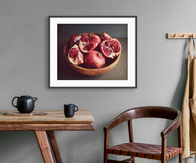 May include: A framed wall art print featuring a wooden bowl filled with several whole and halved pomegranates showing deep red seeds. The artwork is displayed in a dark frame, showcasing a still life composition with rich red and brown tones.