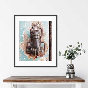 May include: A framed wall art print featuring a weathered, light blue wooden door with a dark, metallic hand-shaped door knocker. The door surface shows significant paint chipping and wood texture. The print is displayed in a thin black frame.