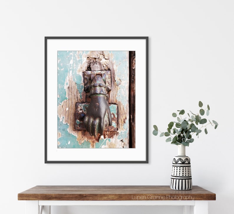 May include: A framed wall art print featuring a weathered, light blue wooden door with a dark, metallic hand-shaped door knocker. The door surface shows significant paint chipping and wood texture. The print is displayed in a thin black frame.
