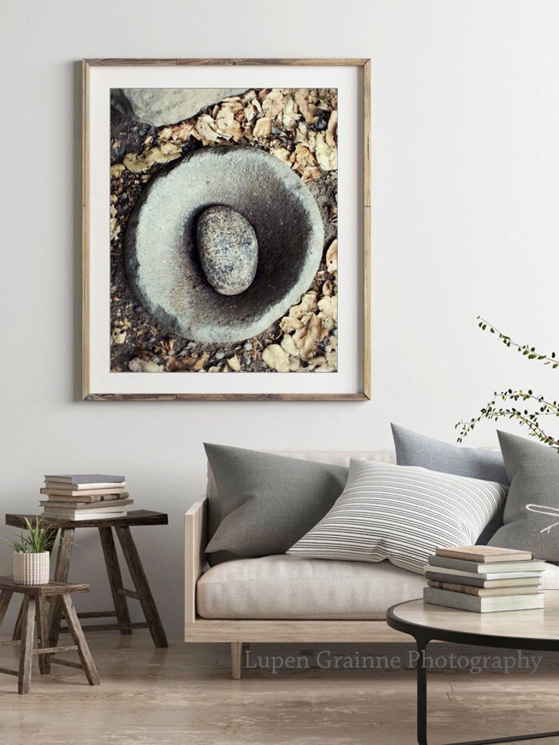 May include: A framed wall art print featuring a high-angle view of a smooth, circular grey stone basin containing a single oval pebble, surrounded by scattered dry brown leaves and dark earth, presented in a light-toned wooden frame.