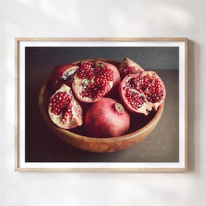 May include: A framed wall art print featuring a wooden bowl filled with several pomegranates. Some fruits are whole with smooth red rinds, while others are cut open to reveal clusters of deep red, glistening arils inside the textured fruit segments.