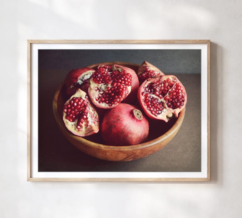 May include: A framed wall art print featuring a wooden bowl filled with several pomegranates. Some fruits are whole with smooth red rinds, while others are cut open to reveal clusters of deep red, glistening arils inside the textured fruit segments.