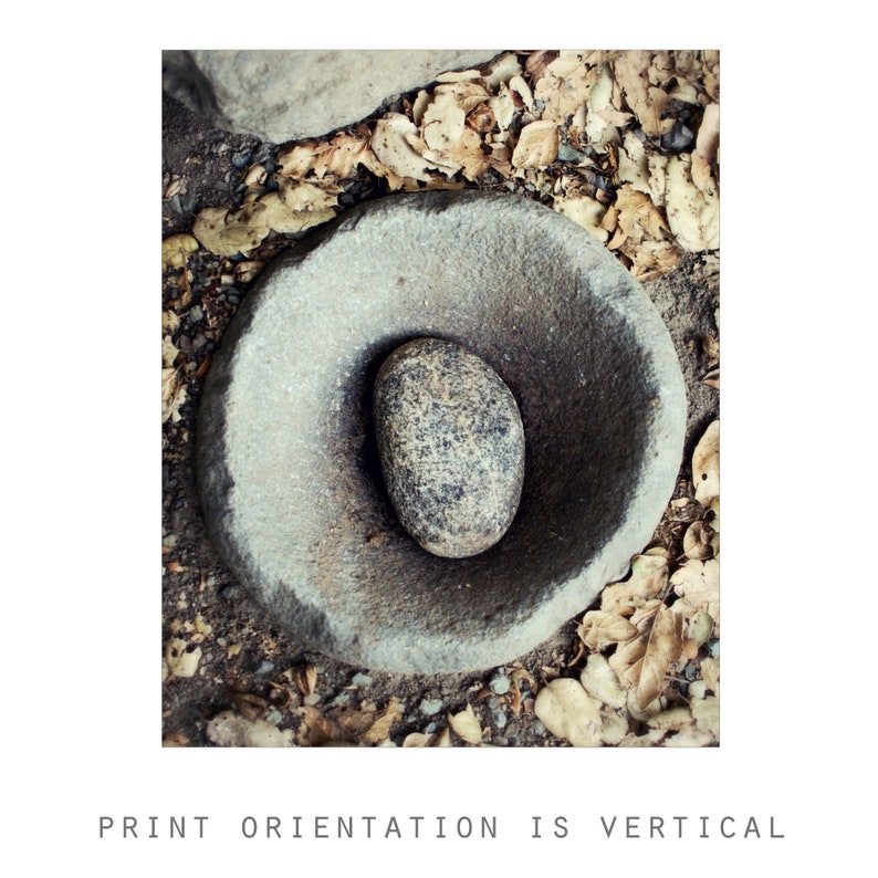 May include: A circular grey stone mortar with a rough, textured surface and a deep, concave centre. Inside the basin rests a smooth, oval-shaped grey stone pestle with dark speckled patterns, creating a functional tool for grinding.