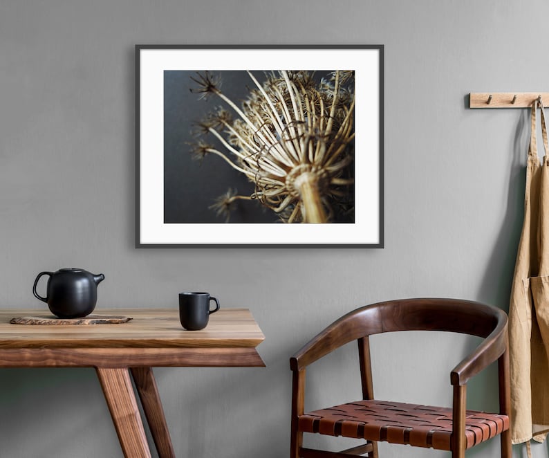 Puede incluir: Una impresi&oacute;n de arte de pared enmarcada que muestra una cabeza de flor de umbela seca de color tostado sobre un fondo oscuro. La obra est&aacute; en un marco negro fino, colocada sobre una mesa de madera con una tetera y una taza negras.