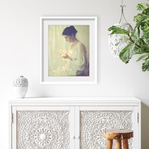 May include: A framed art print featuring a woman holding glowing lights. The artwork has a soft, vintage aesthetic with a white frame. The image is displayed on a white cabinet with carved floral designs, alongside a decorative vase and a small wooden stool.