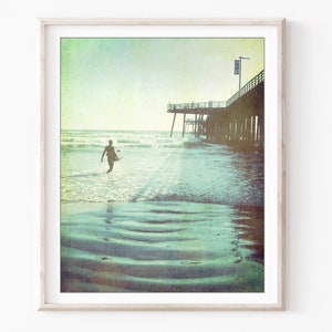 Photograph of a surfer walking out to the ocean next to a wooden pier at Pismo beach California, with a vintage style. Fine art photography print.